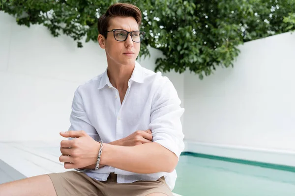 Man with silver bracelet and eyeglasses sitting near pool and looking away — Stockfoto