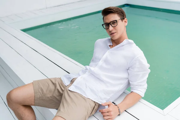 Thoughtful man in summer clothes and eyeglasses looking at camera near pool — Stockfoto