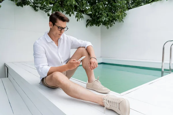 Full length of man in summer clothes and eyeglasses messaging on smartphone near pool — Stock Photo