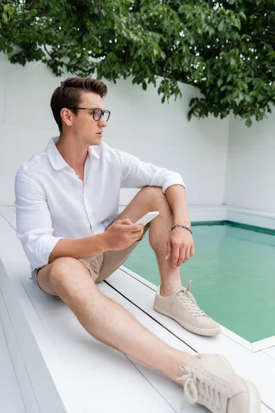 Full length of man in sneakers sitting near pool with mobile phone — Stockfoto