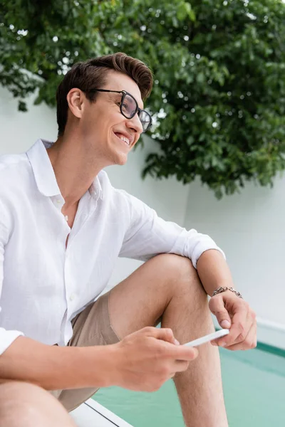 Радостный мужчина в белой рубашке и очках держит смартфон и смотрит в сторону бассейна — стоковое фото