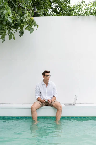 Man in white shirt sitting near pool and laptop while holding mobile phone — Stock Photo