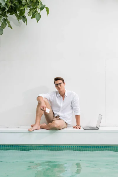 Повнометражний босоніж чоловік зі смартфоном, що сидить біля басейну біля ноутбука — стокове фото