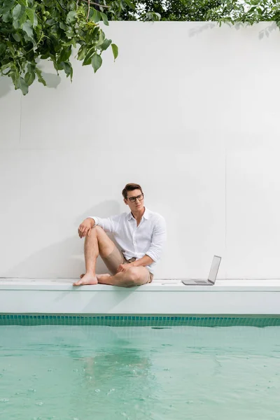 Full length of barefoot man in eyeglasses sitting at poolside near white wall and laptop — Stock Photo