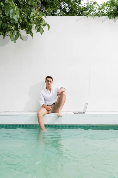Freelancer in eyeglasses sitting at white wall near pool and laptop — Stock Photo