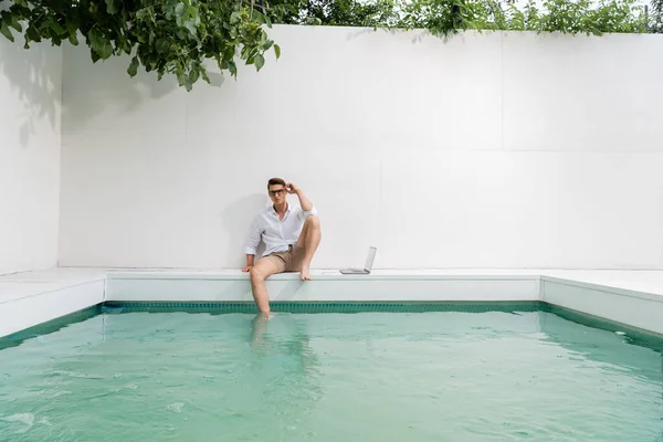 Freelances sitting at poolside near laptop and white wall under green branches - foto de stock