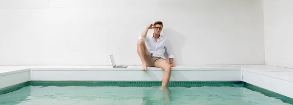 Homem de camisa branca sentado à beira da piscina perto do laptop e ajustando óculos, banner — Fotografia de Stock