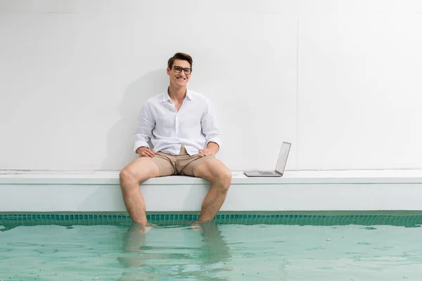 Pleased man sitting at poolside with feet and water near white wall and laptop - foto de stock