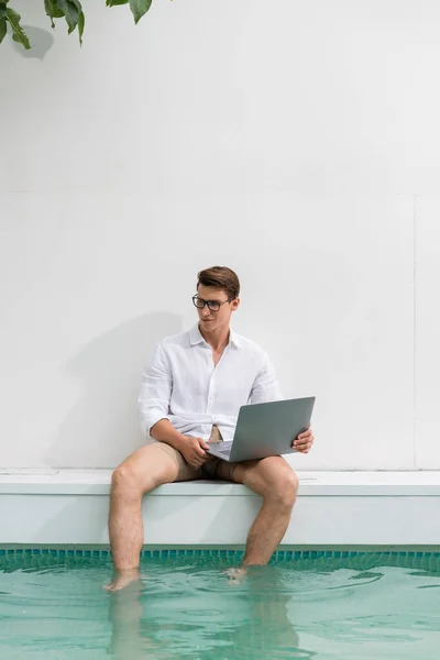 Freelancer in eyeglasses holding laptop while sitting with feet in pool — Stock Photo