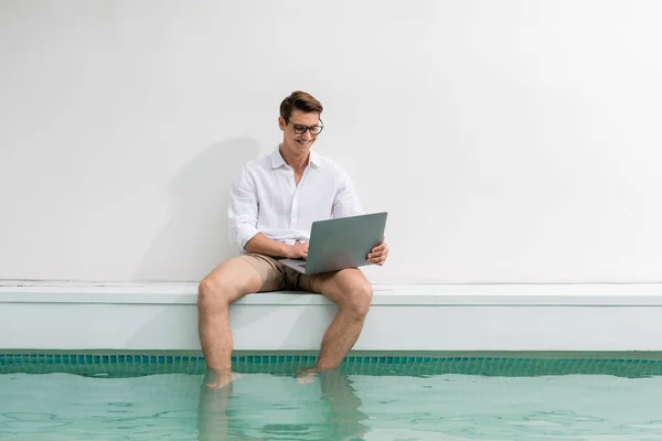 Homem alegre sentado na piscina com os pés na água e usando laptop — Fotografia de Stock