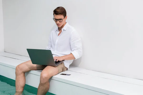 Man in eyeglasses tying on laptop while sitting at poolside near mobile phone — Fotografia de Stock