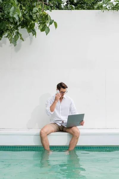 Man sitting with feet in pool and talking on mobile phone near laptop — Stock Photo