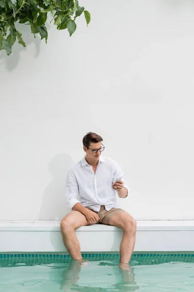 Smiling man sitting near pool at white wall and looking at smartphone — Stock Photo