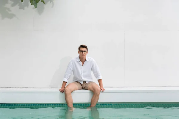 Man in white shirt and eyeglasses sitting with feet in pool by white wall — Stock Photo