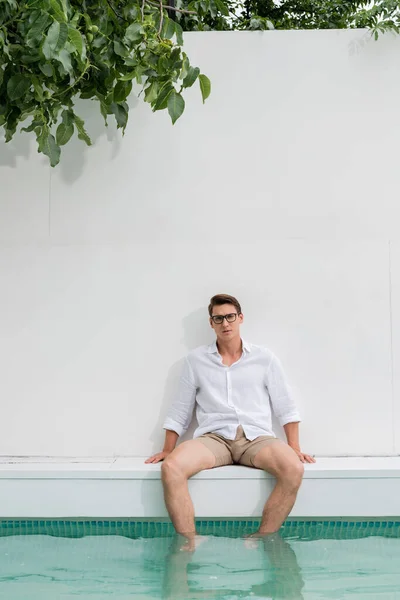 Man in shirt and eyeglasses sitting near pool by white wall — Stock Photo