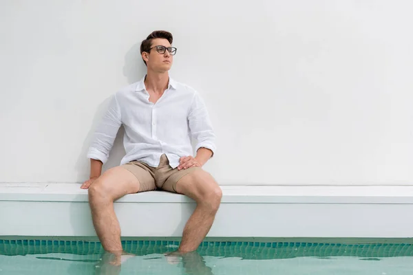 Мрійливий чоловік в окулярах дивиться в сторону, сидячи з ногами в басейні — стокове фото