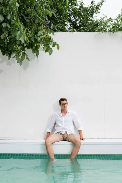 Smiling man sitting near pool by white wall under green branches - foto de stock