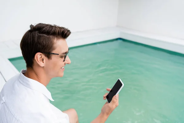 Усміхнений чоловік має відеодзвінок на смартфон біля басейну з бірюзовою водою — стокове фото