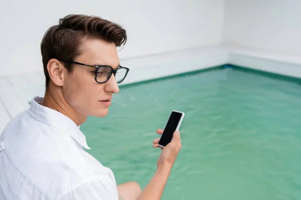 Mann mit Brille hält Smartphone mit leerem Bildschirm nahe verschwommenem Pool — Stockfoto