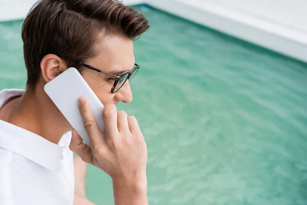 Uomo positivo negli occhiali che parla sul cellulare vicino alla piscina con acqua turchese — Foto stock