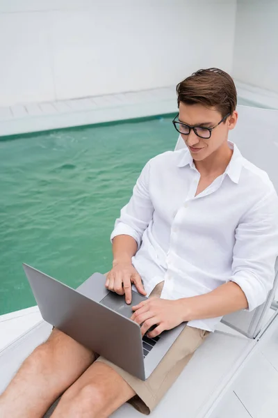 High angle view of freelancer typing on laptop in deck chair near pool with turquoise water — Photo de stock