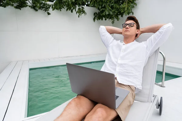 Man with closed eyes relaxing near pool while sitting in deck chair with laptop — Photo de stock