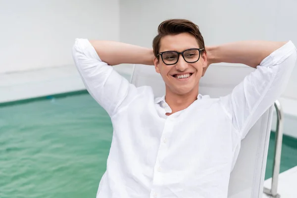 Fröhlicher Mann mit Brille, der wegschaut, während er mit den Händen hinter dem Kopf in der Nähe des Pools ruht — Stockfoto