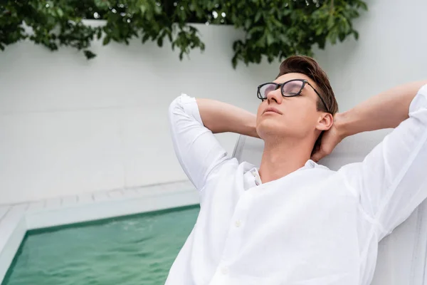 Homem em óculos relaxante com os olhos fechados e as mãos atrás da cabeça perto da piscina — Fotografia de Stock