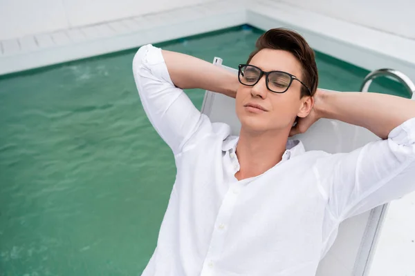 Чоловік у білій сорочці та окулярах розслабляється із закритими очима та руками за головою біля басейну — стокове фото