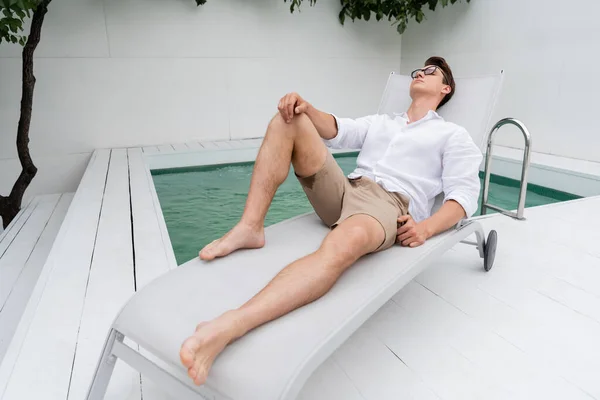 Full length of barefoot man in summer clothes resting on deck chair at poolside — Photo de stock