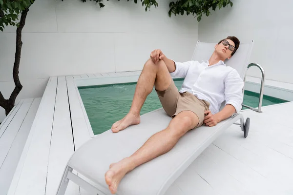 Barefoot man in shorts and eyeglasses lying on deck chair near pool — Photo de stock