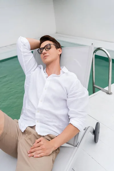 Man in white shirt and eyeglasses resting in deck chair near pool and looking at camera — Stockfoto