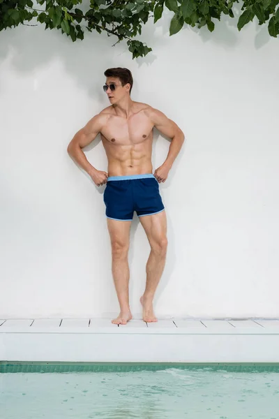 Full length of man in sunglasses standing with hands on hips by white wall near pool — Stock Photo