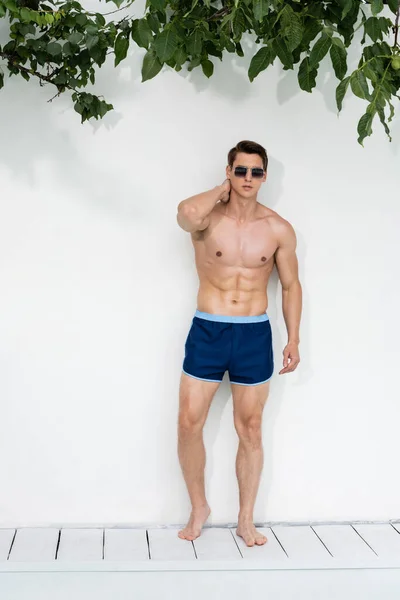 Athletischer Mann in Badehose und Sonnenbrille posiert in der Nähe der weißen Wand — Stockfoto