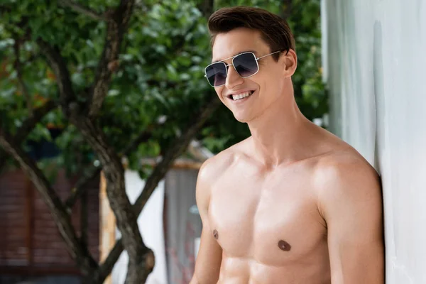 Homem sem camisa positivo em óculos de sol sorrindo perto da parede ao ar livre — Fotografia de Stock