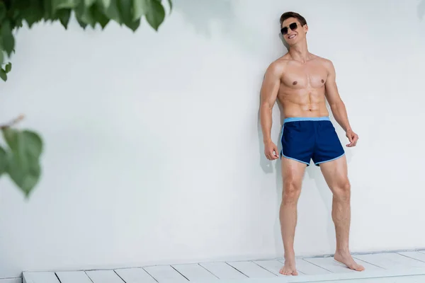 Full length of smiling sportive man in sunglasses and swimming trunks near white wall — Stock Photo