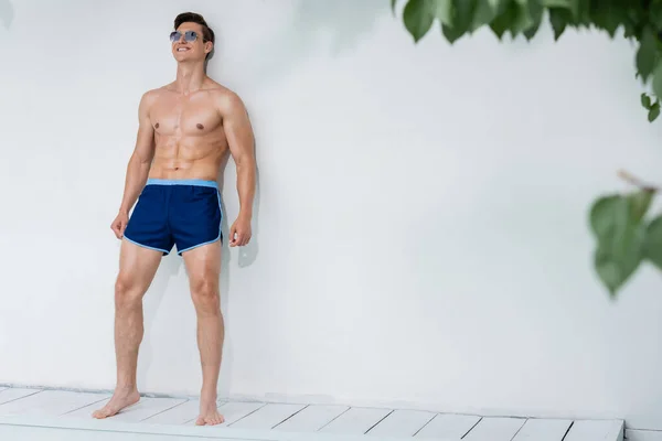 Comprimento total do homem com o corpo muscular em pé pela parede branca em calções de banho e óculos de sol — Fotografia de Stock