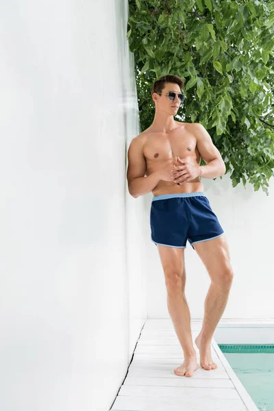 Full length of sportive man in sunglasses and swimming trunks posing by white wall at poolside — Foto stock