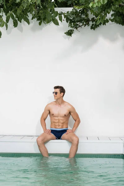 Homem sorridente em óculos de sol e calções de banho sentado perto da piscina e olhando para longe — Fotografia de Stock