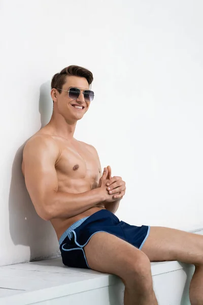 Cheerful athletic man in sunglasses and swimwear sitting by white wall - foto de stock