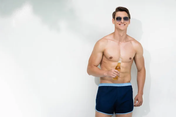 Happy man holding bottle of beer while standing in sunglasses and swimwear near white wall — Fotografia de Stock