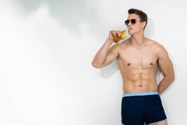 Shirtless man in sunglasses standing near white wall with hand behind back and drinking beer — Photo de stock