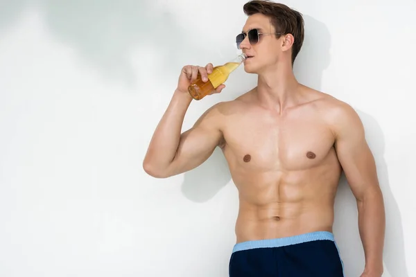 Sportive man in sunglasses drinking beer while standing by white wall — стоковое фото