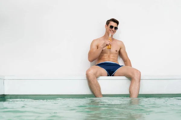 Happy man in sunglasses and swimming trunks drinking beer near pool by white wall — Fotografia de Stock