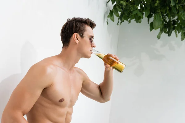 Shirtless and sportive man in sunglasses drinking beer near white wall — Stock Photo