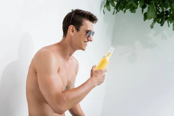 Cheerful shirtless man in sunglasses looking at bottle of beer outdoors — Stock Photo