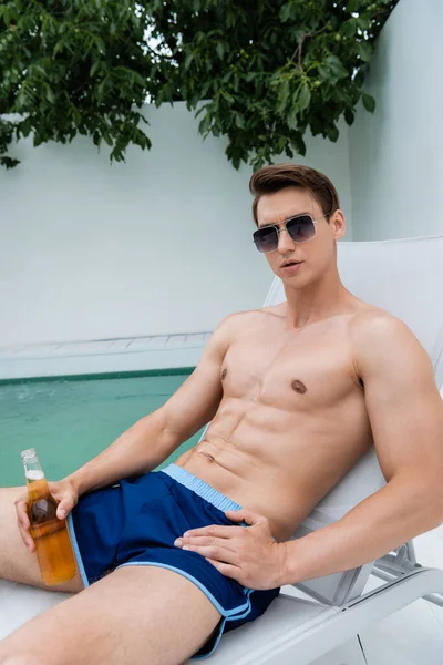 Мужчина без рубашки в солнечных очках сидит в шезлонге с бутылкой пива — стоковое фото