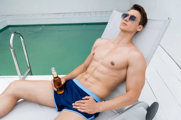 Man with bottle of beer relaxing in deck chair in sunglasses and swimming trunks — стоковое фото