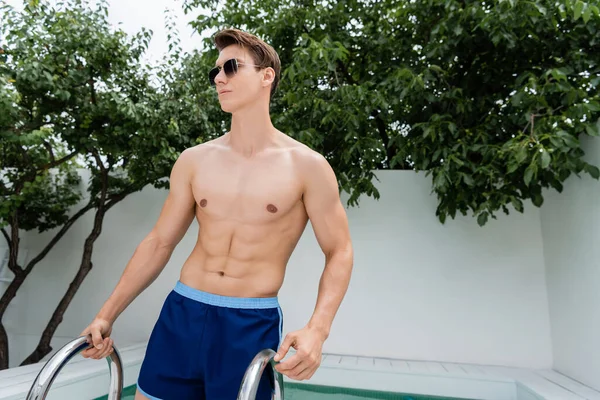 Shirtless man in sunglasses looking away near pool ladder — Photo de stock