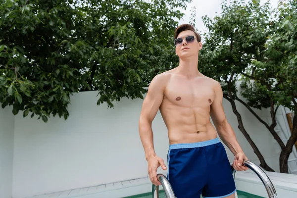 Sportive man in sunglasses standing at pool ladder and looking away - foto de stock